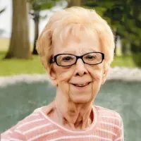 Smiling woman with short blonde hair and glasses, wearing a pink striped shirt outdoors by a pond.