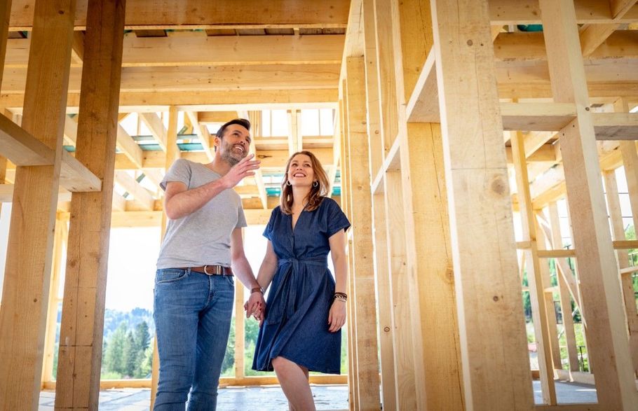 Two people examining a wooden house frame at a construction site, with sunlight streaming through the beams.