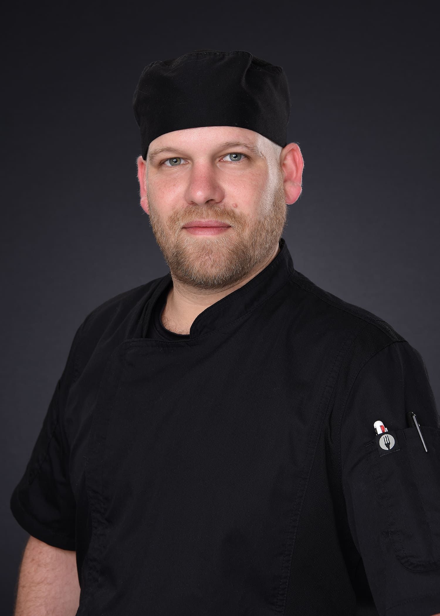 A man with a beard is wearing a black chef 's hat and a black shirt.