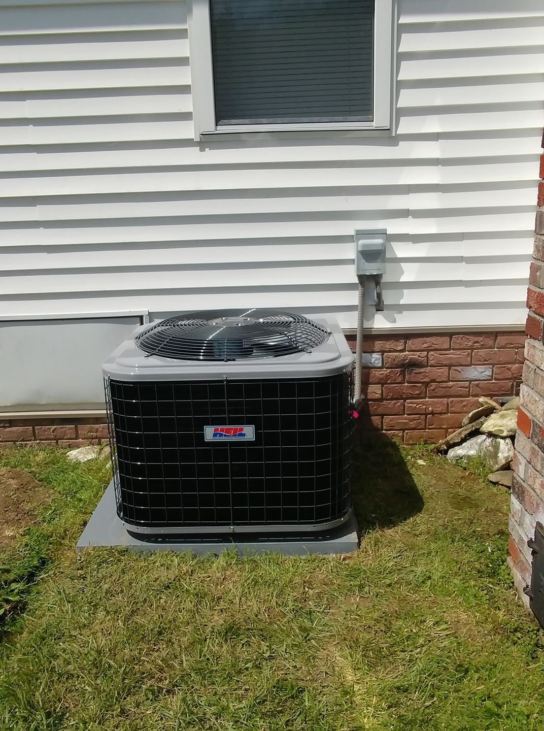 A black air conditioner is sitting in the grass in front of a white house.