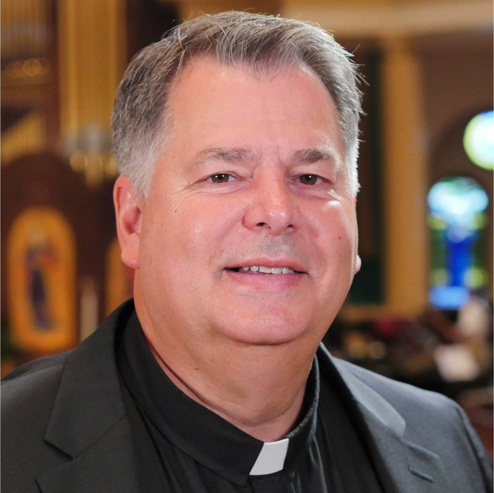 Clergyman wearing a clerical collar, smiling, set in a church.