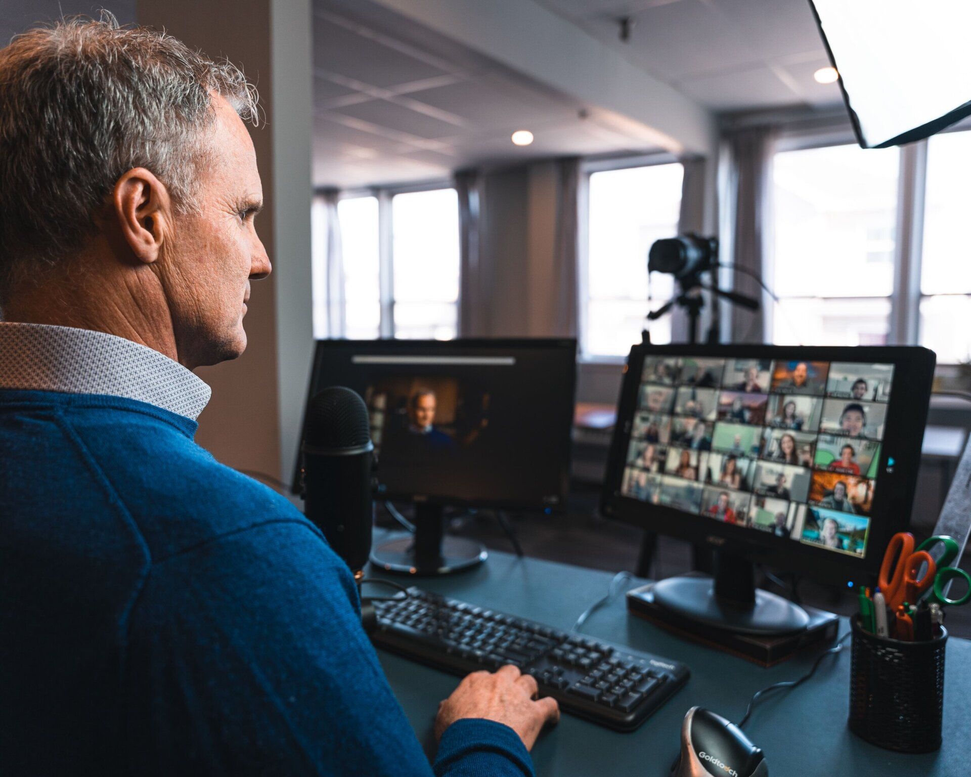 Man in blue sweater at desk, participating in a video conference with multiple participants on a screen.