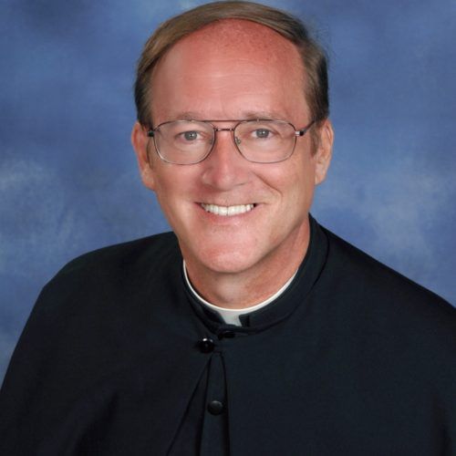 Man wearing glasses and clerical collar, smiling, against a blue background.