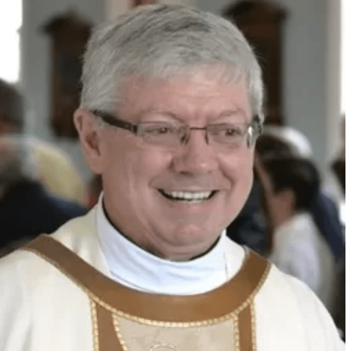 Clergyman smiling, wearing glasses and a white vestment with gold trim; blurred background.