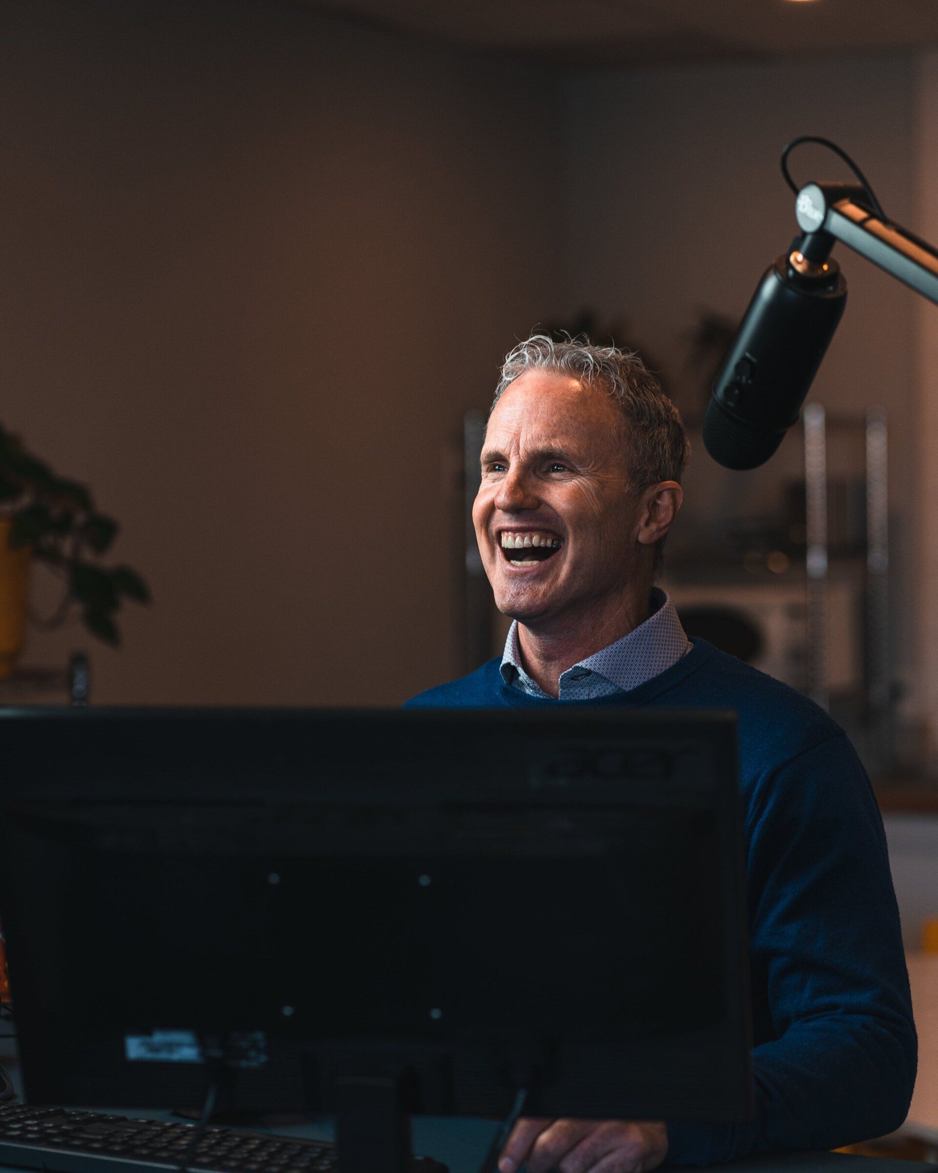 Man with graying hair laughs, speaking into a microphone. Dark blue sweater, computer screen, and office setting.