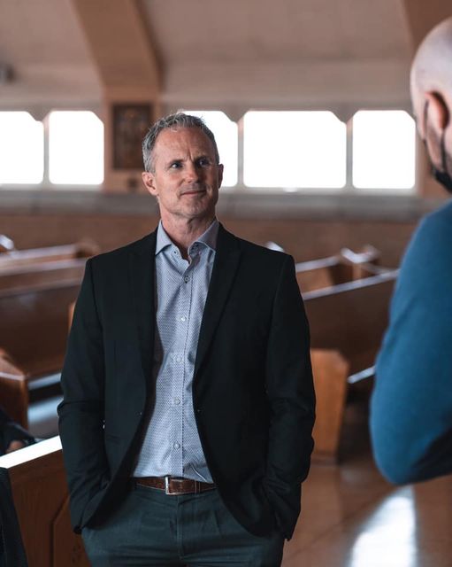 Man in a blazer and open-collared shirt, standing inside a church, looking toward another person.