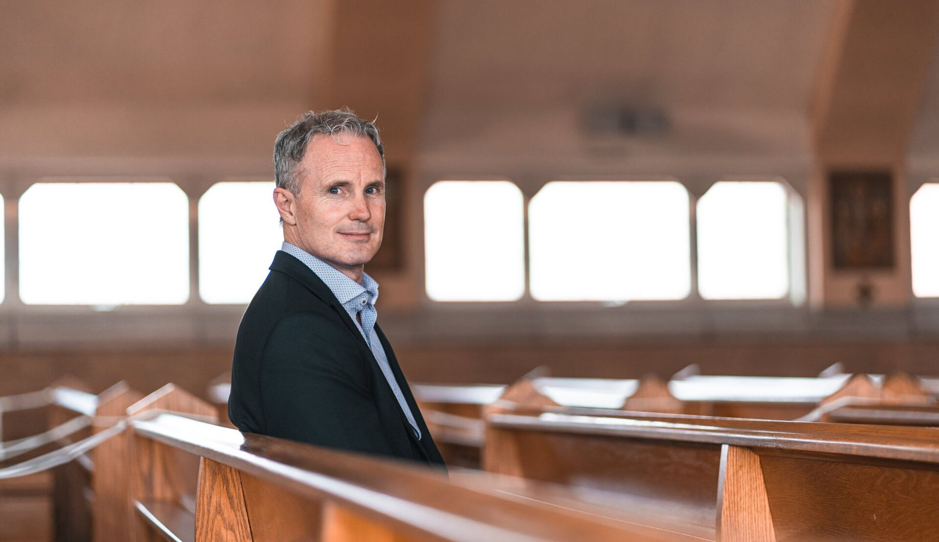 Man in a blazer sits in a church pew, looking at the camera.