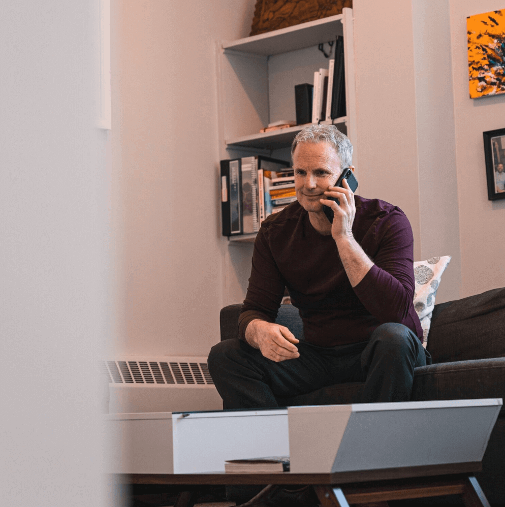 Man with gray hair sits on a couch, talking on a phone near a laptop, in a living room.