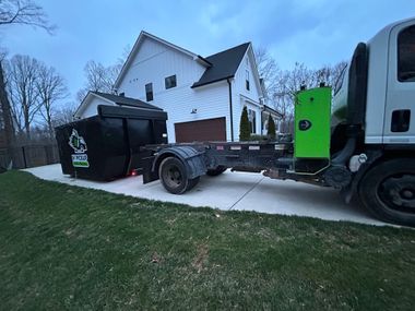 Truck parked beside a white house, with a black dumpster and green equipment box on a concrete driveway.