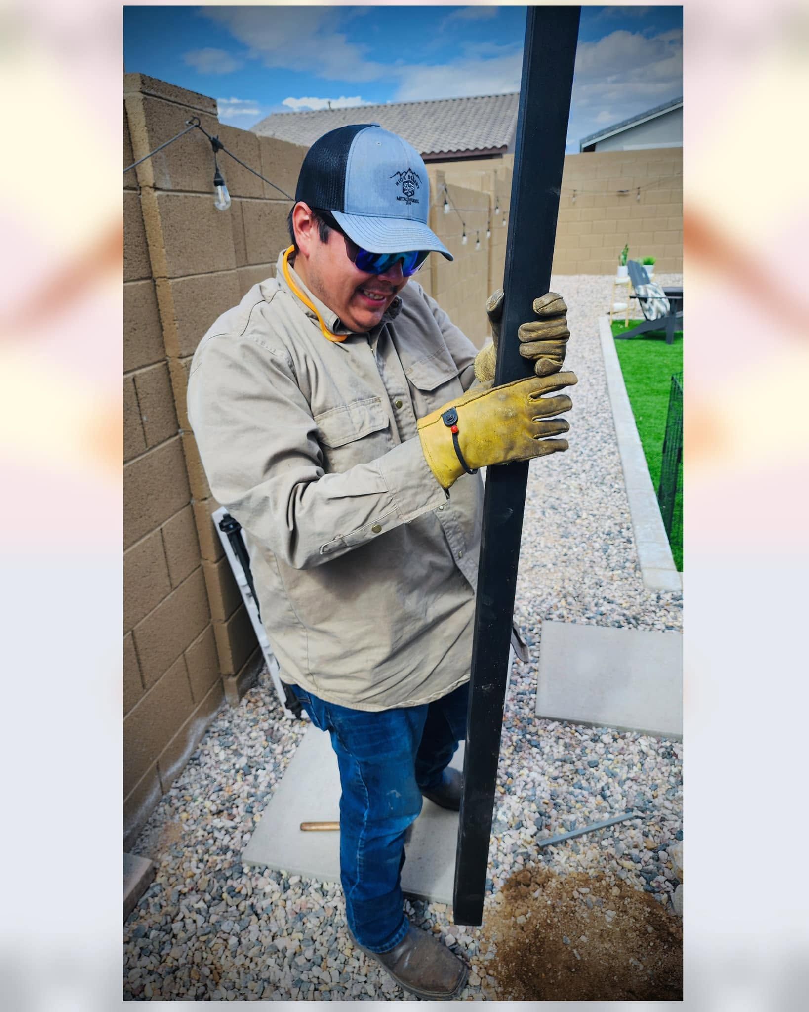 Worker in beige jacket and blue cap installing a black post beside a gravel walkway and stone wall