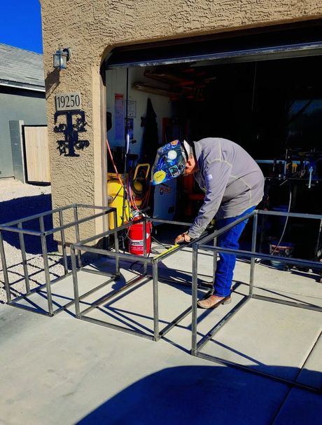 Person welding a black metal gate frame in a garage doorway, wearing a mask and blue pants.