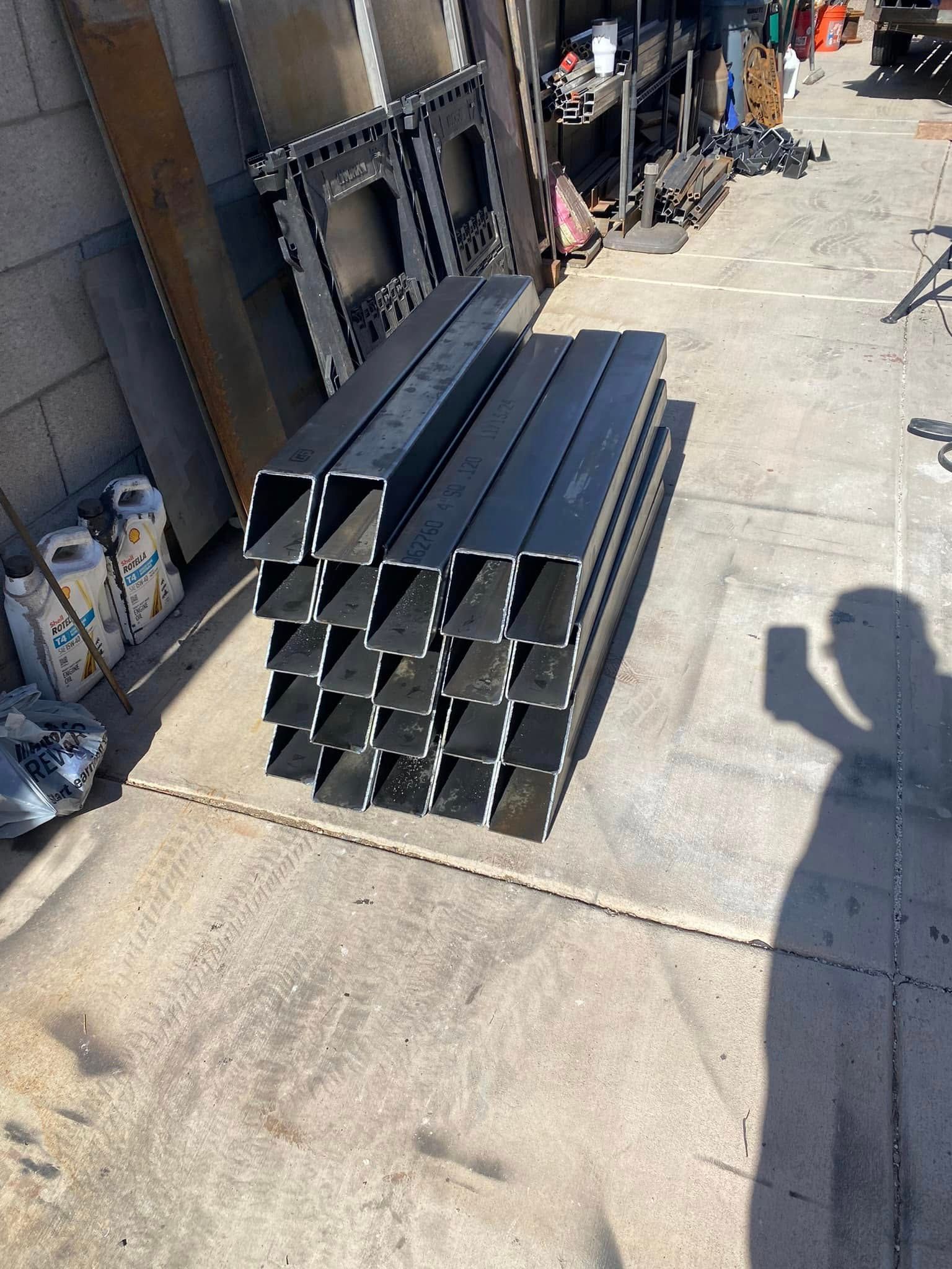 Stacked black metal beams on a concrete sidewalk beside a building, with a person’s shadow in the foreground
