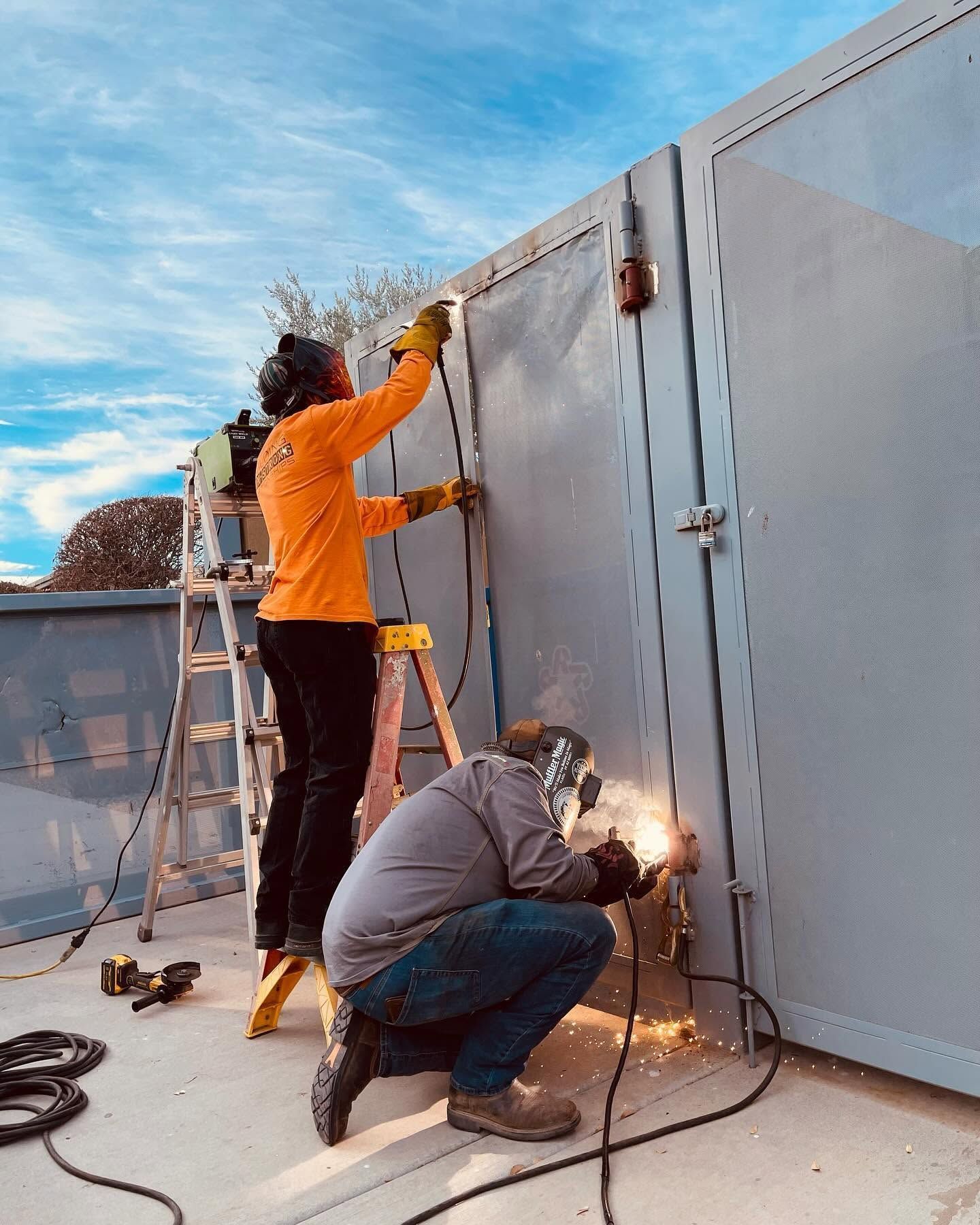 Two workers welding a large metal wall outdoors, one standing on a ladder and one crouching with sparks flying