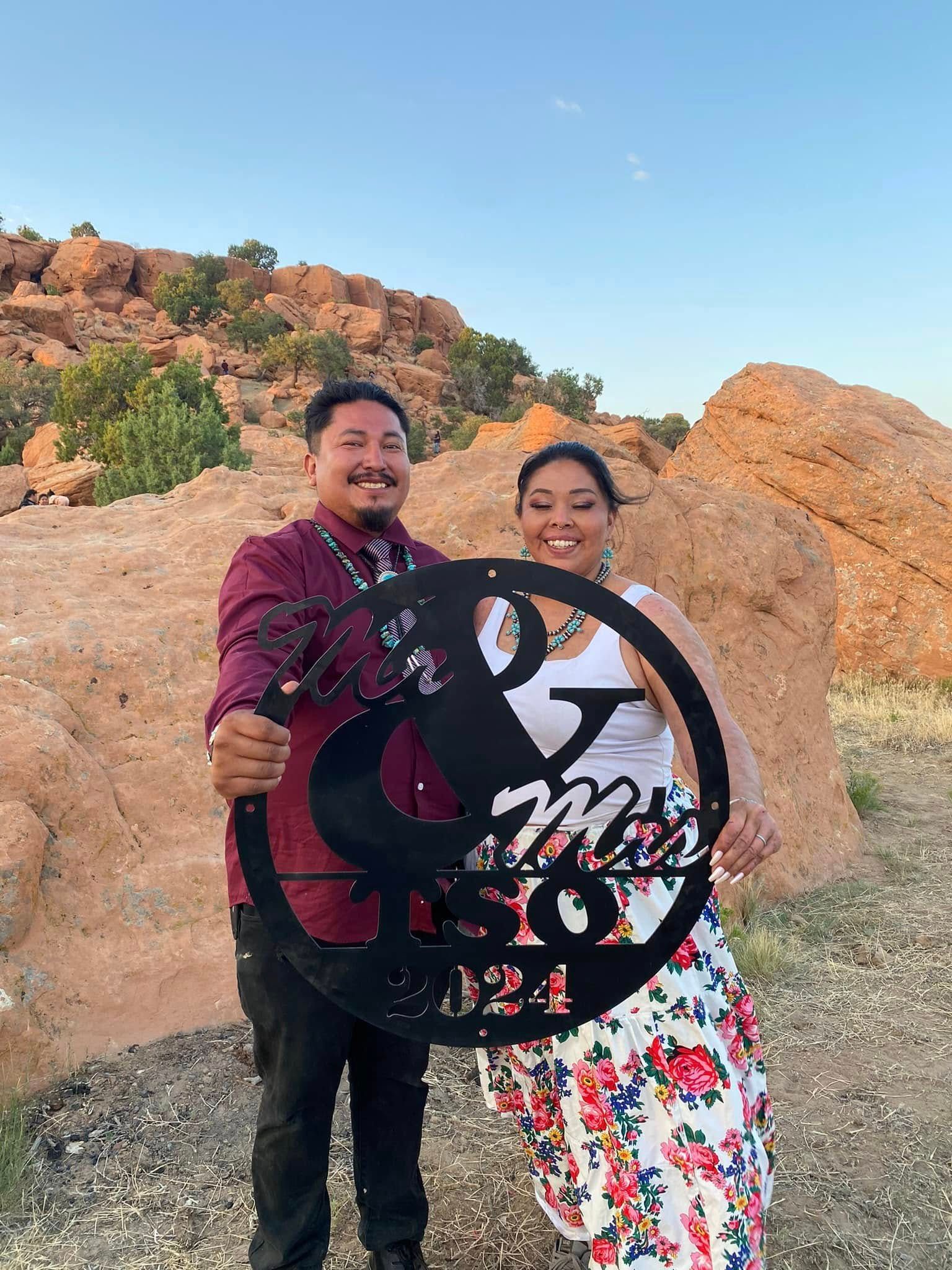 Two people smiling outdoors at sunset, holding a large black circular sign in front of rocky hills.