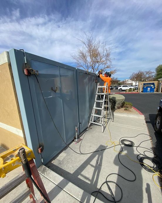 Worker on ladder repairing a large blue metal gate beside a sidewalk