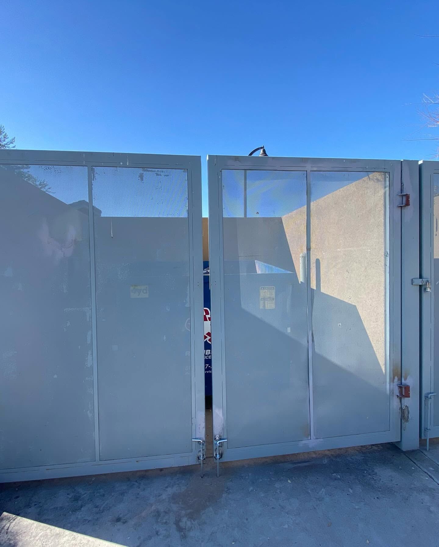 White metal gate doors partially open under a bright blue sky, with a shadow cast across the panels.