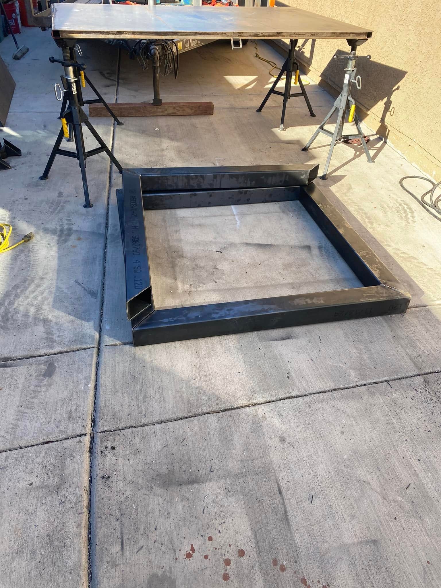 Black square frame on pavement under a workbench, with sawhorses in a workshop area.