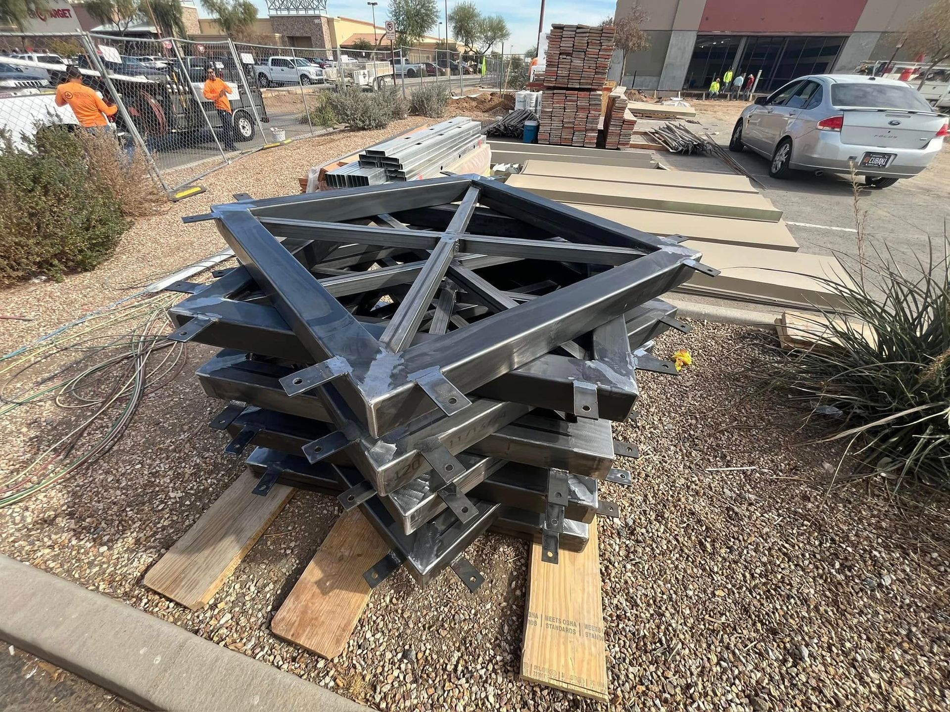 Stacked black metal truss frames on a gravel sidewalk beside a street and parked cars.