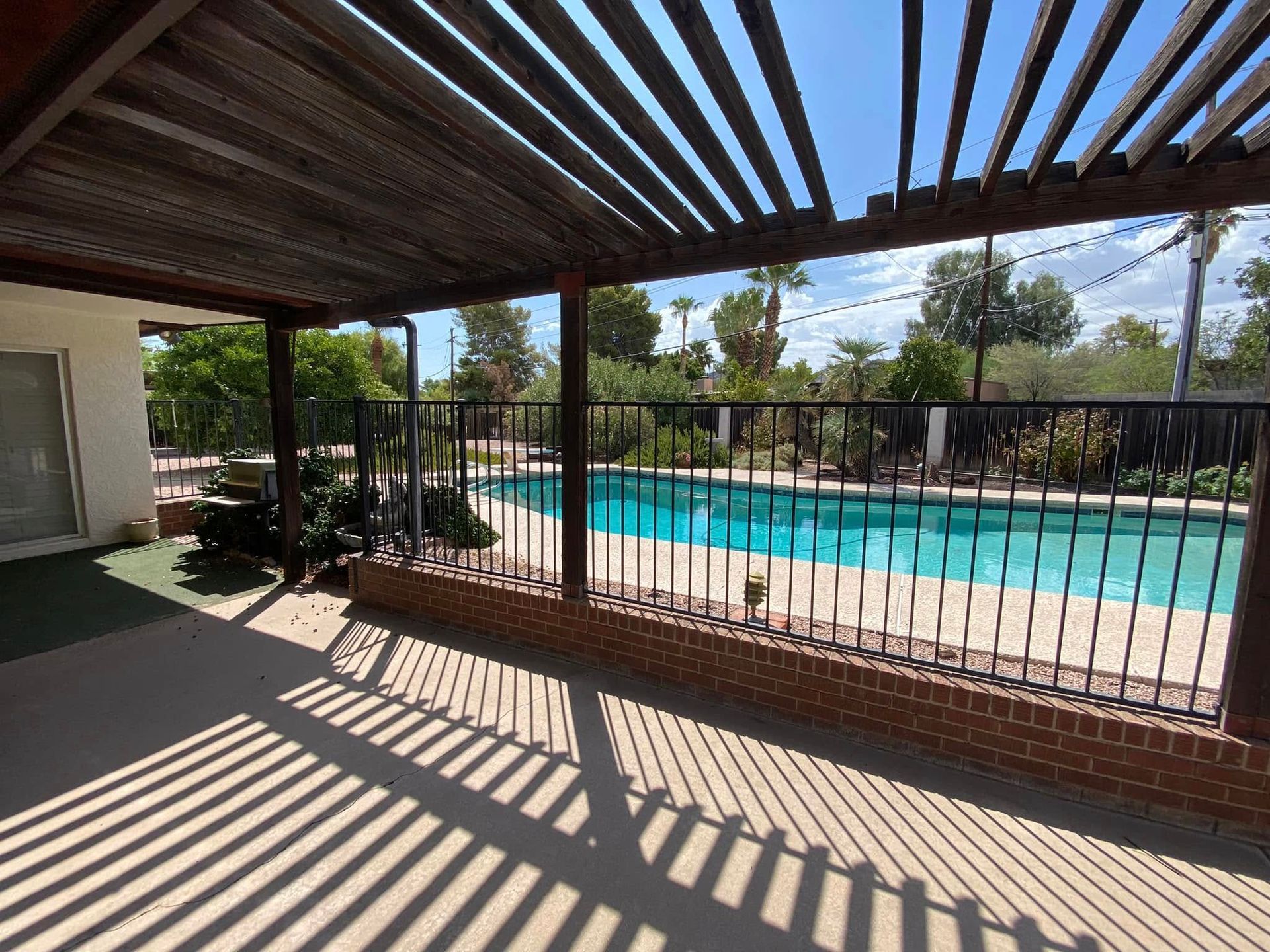 Shaded patio overlooking a swimming pool with striped shadows and a metal fence