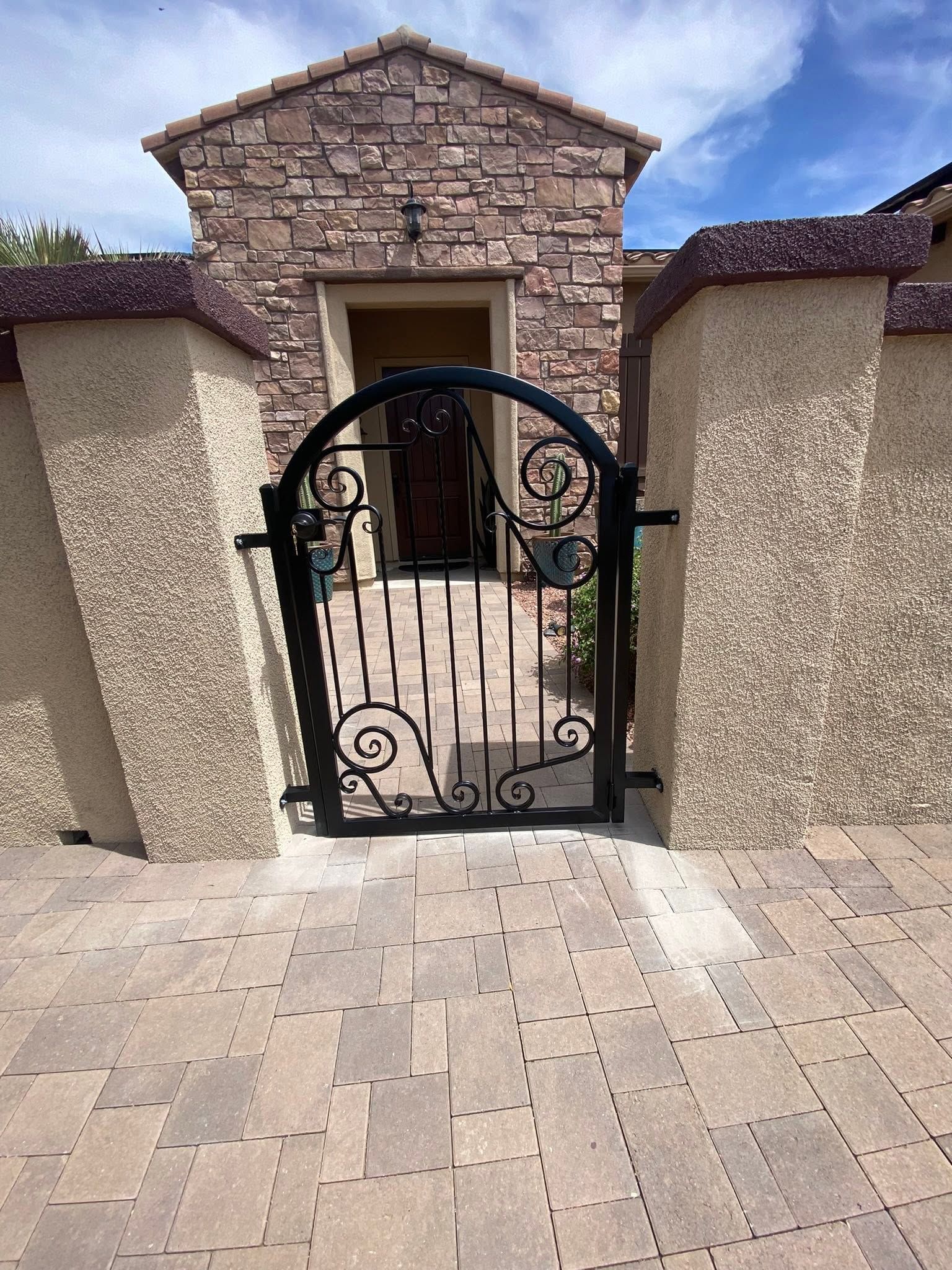 Stone entry gate with black wrought-iron gate and stucco walls under a blue sky