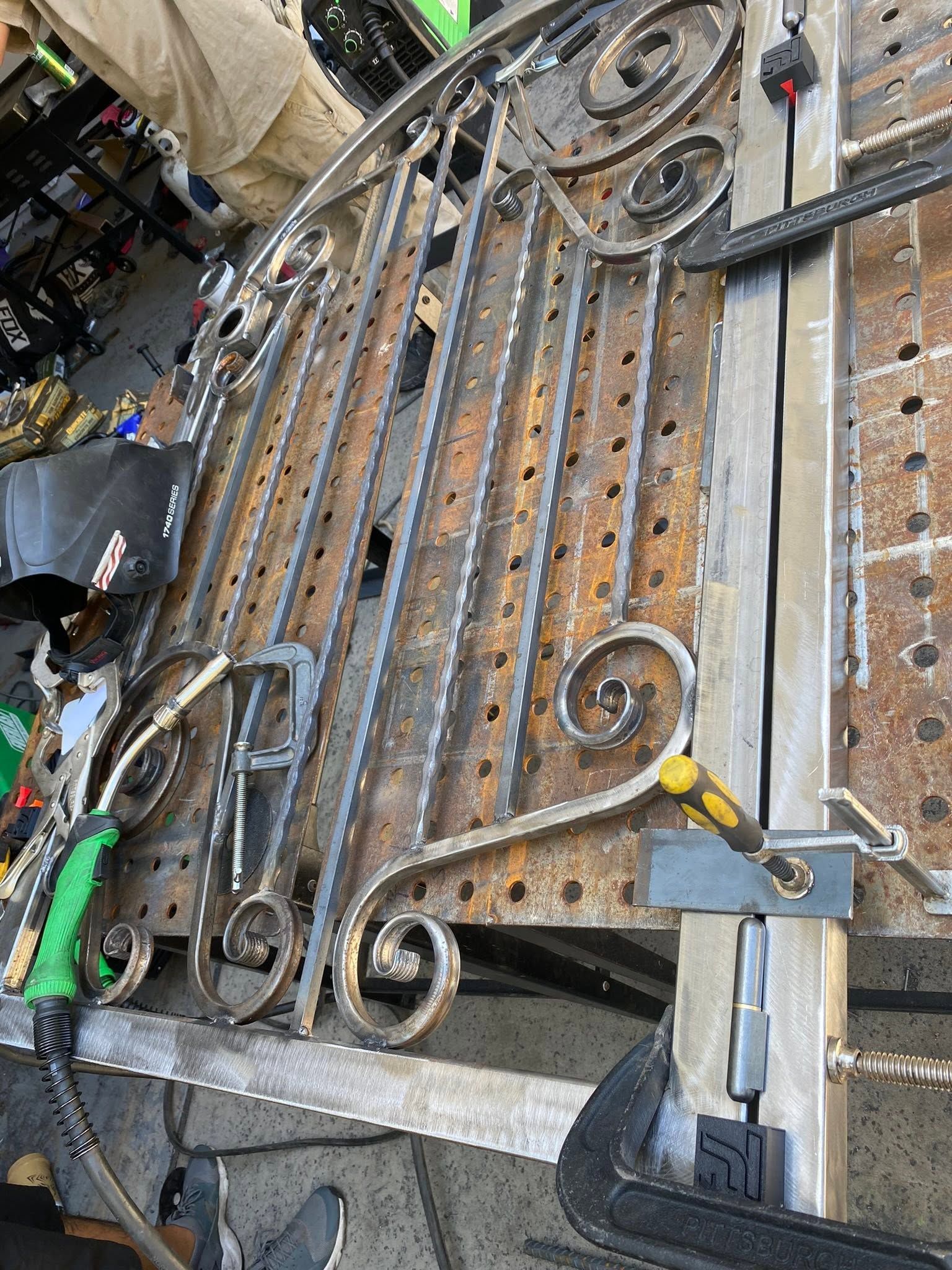 Ornate black iron gate panel leaning in a workshop, with clamps and tools nearby.