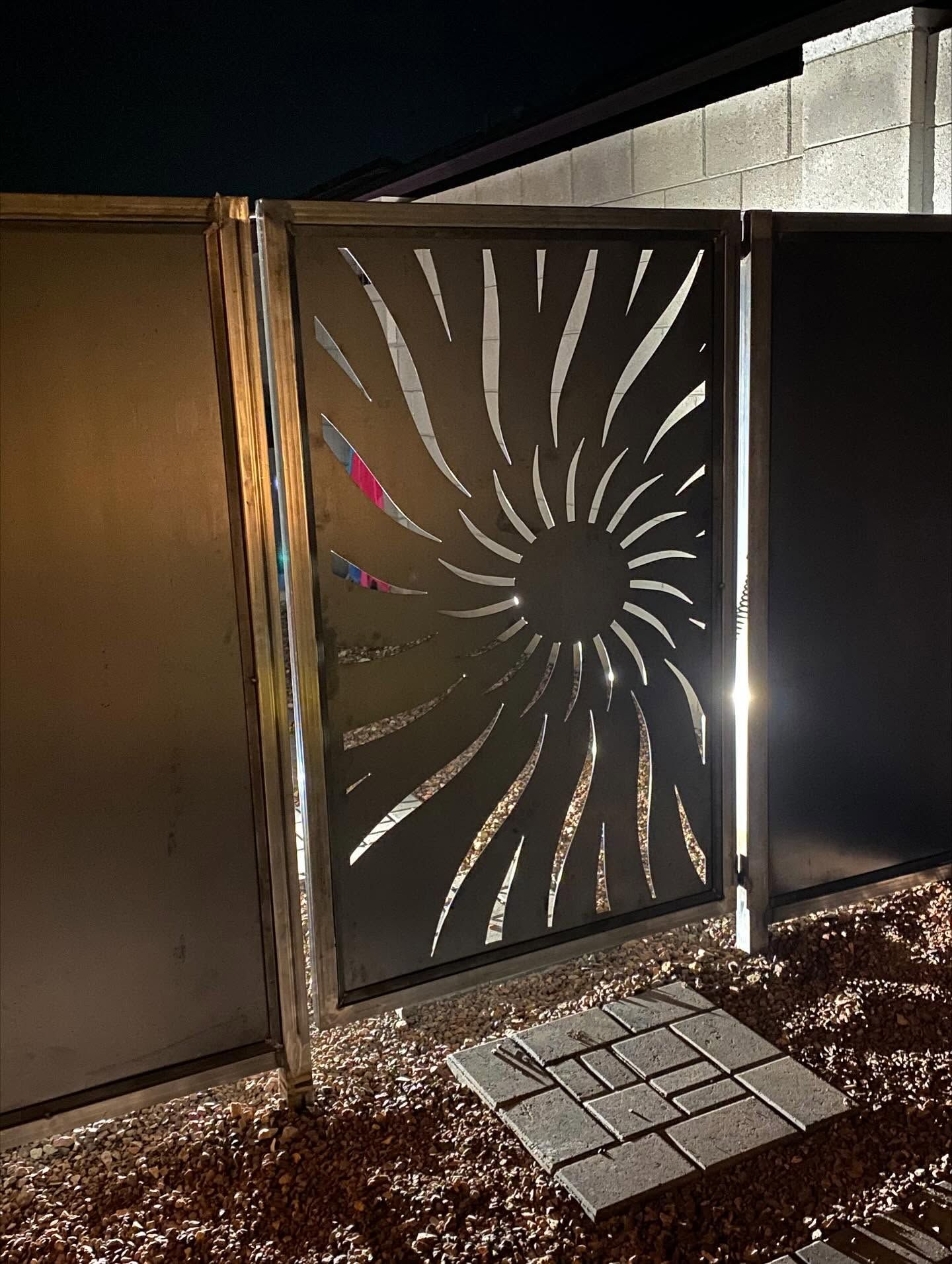 Metal gate with sunburst cutout panels beside a paved walkway at night