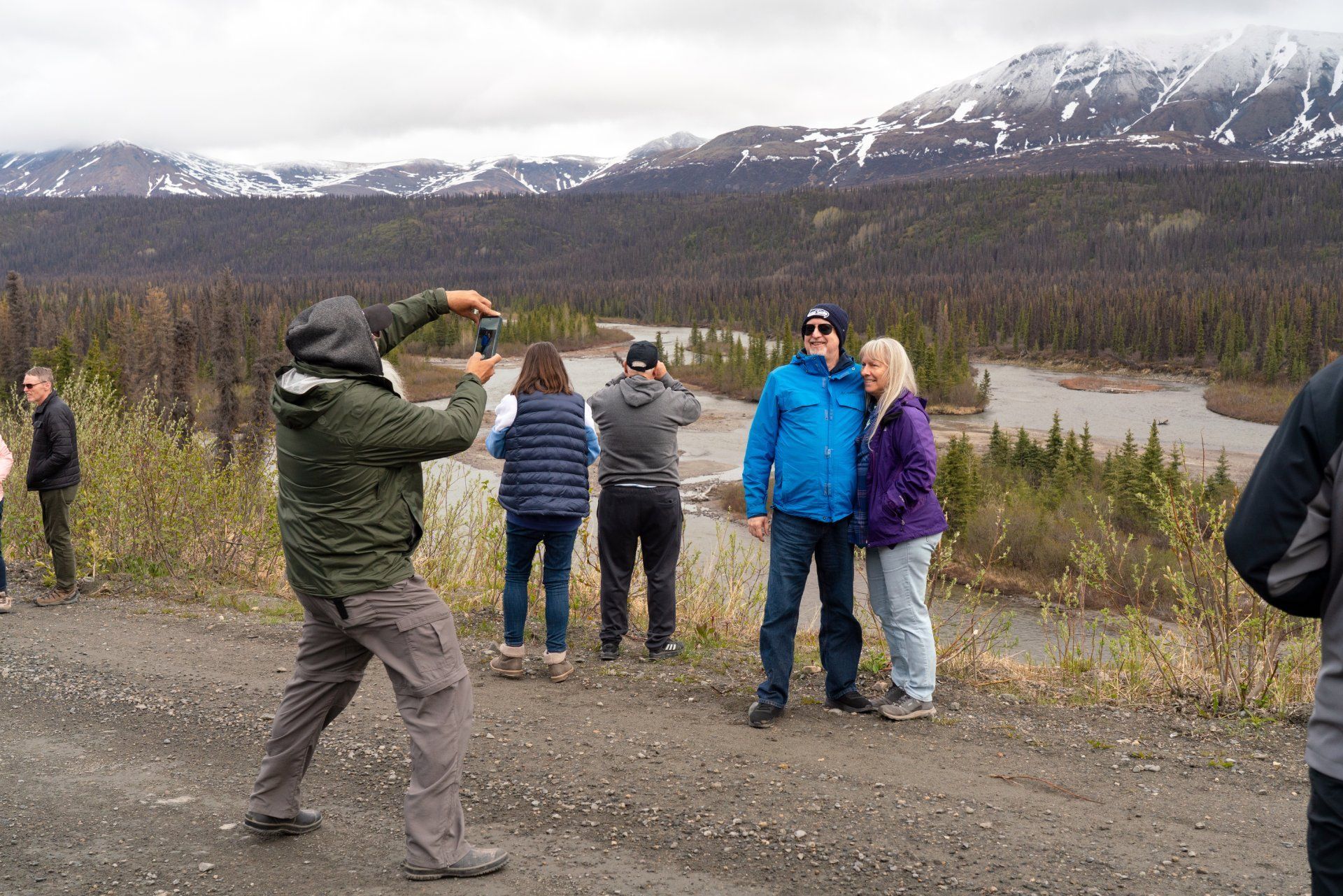 Tourists taking photos at a mountain overlook with snow-capped peaks and a lake in the background