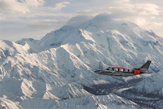 Small plane flying over snow-covered mountains under a cloudy sky
