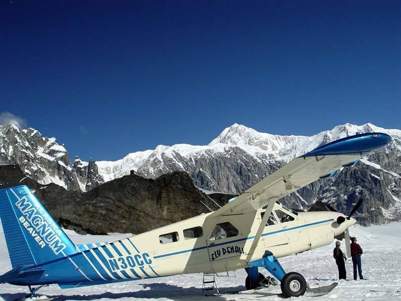 Small blue-and-white plane on snowy mountain airstrip with two people and snow-covered peaks in the background