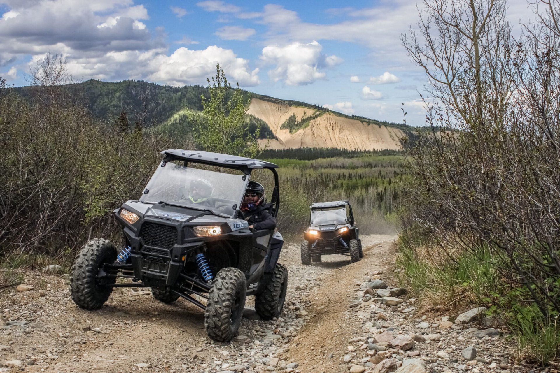 Two off-road ATVs driving on a rocky trail with mountains and trees in the background