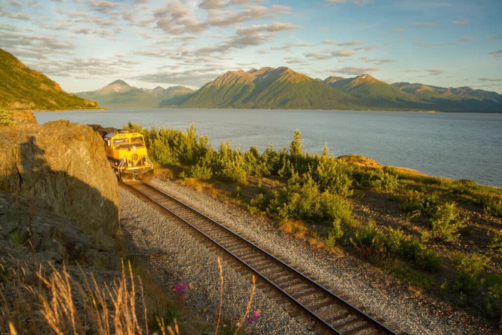 Train on tracks beside a lake, with mountains and a golden sunset sky in the background