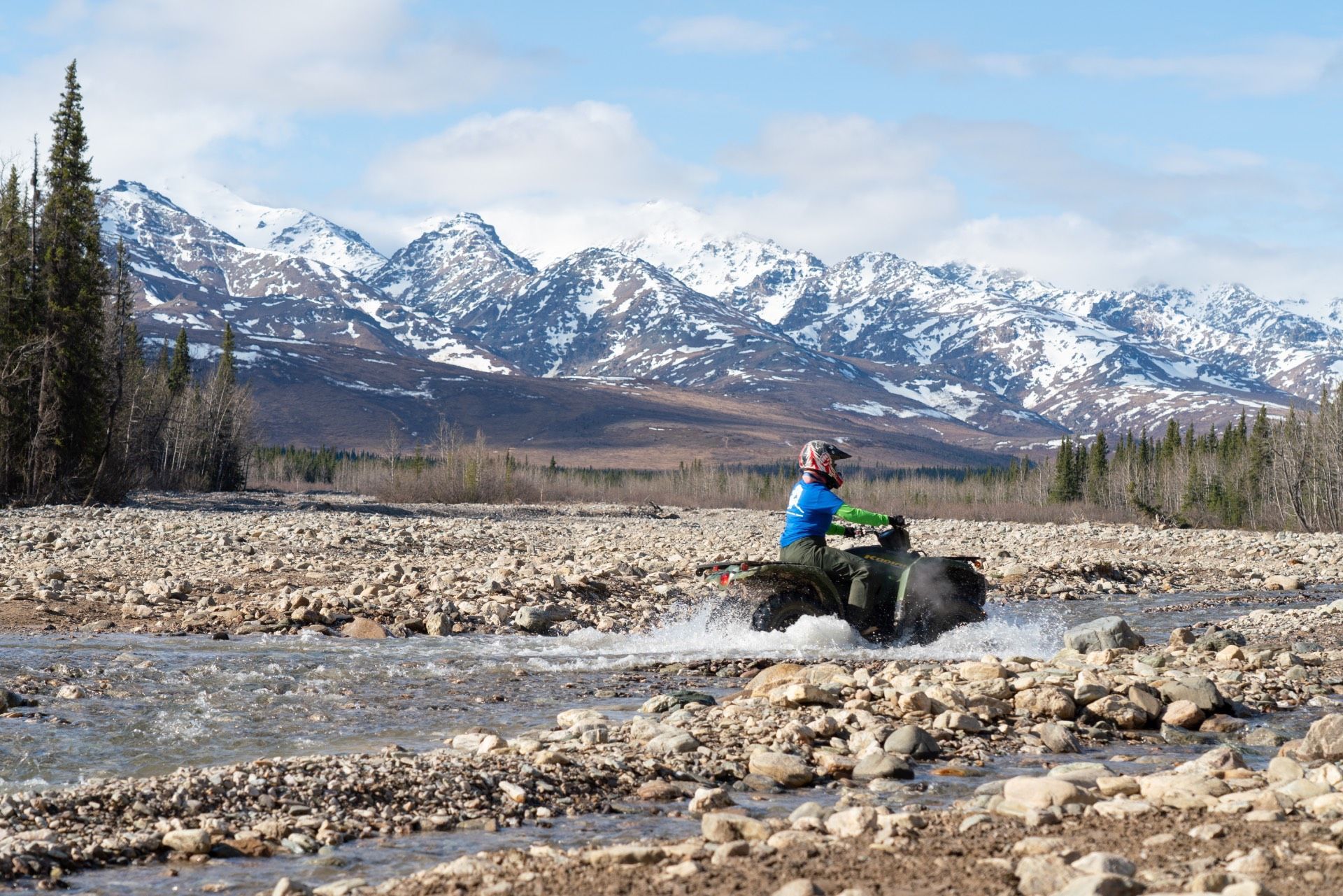 Person on an ATV crossing a rocky river with snow-covered mountains in the background