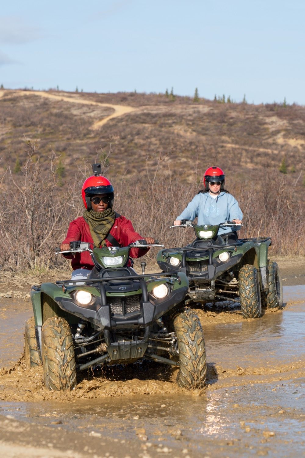Two ATV riders in helmets driving through muddy water on a rural trail