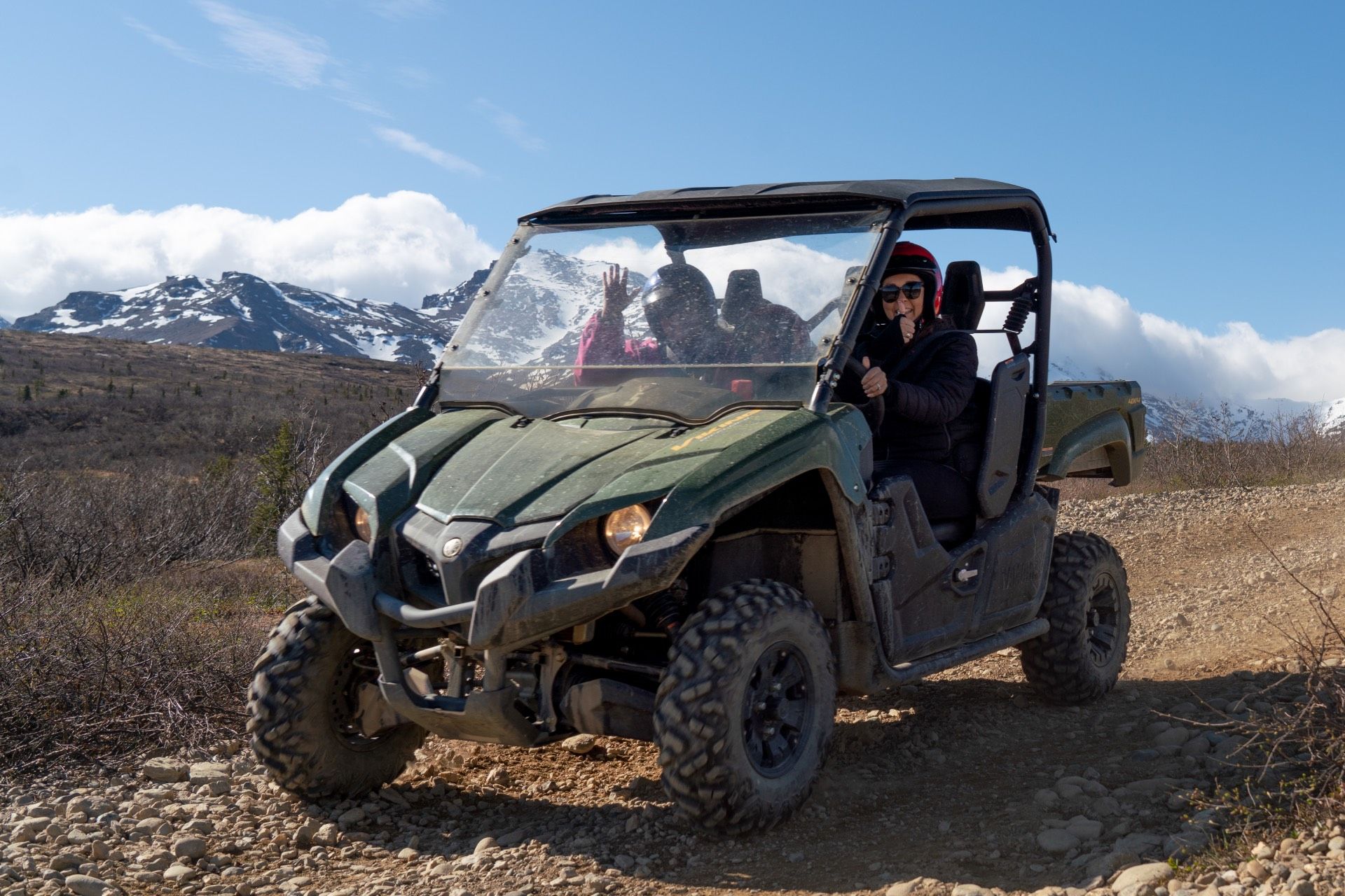 Green off-road UTV with passengers on a dirt trail, snowy mountains in the background