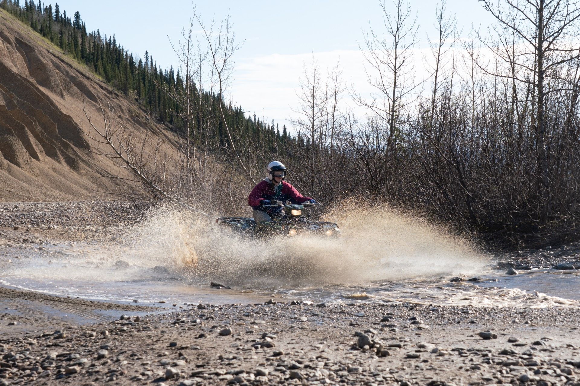 ATV rider splashes through a muddy river in a rocky forest clearing