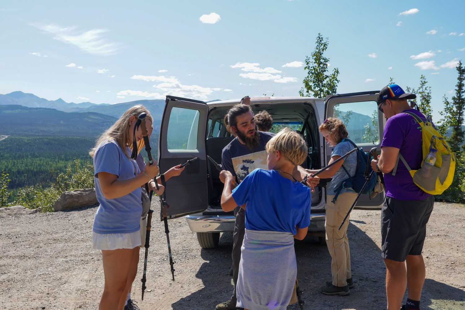 People gathering by a van at a scenic mountain overlook, holding gear in bright sunlight