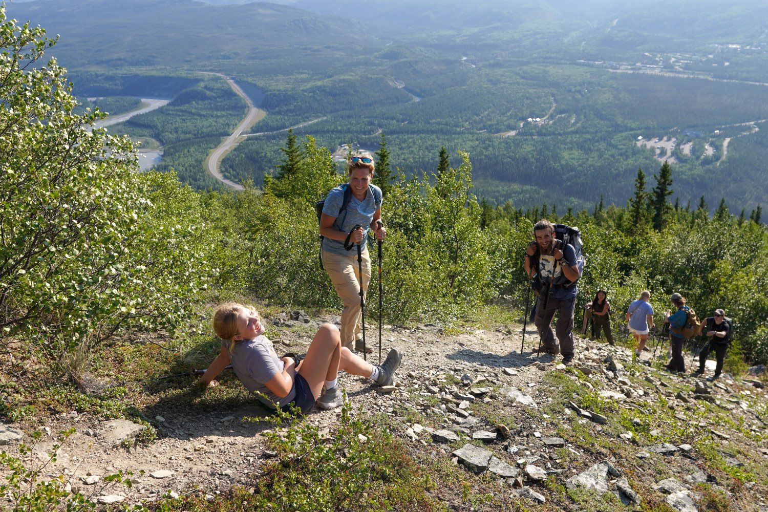 Hikers resting on a rocky mountain trail overlooking a green valley and winding road