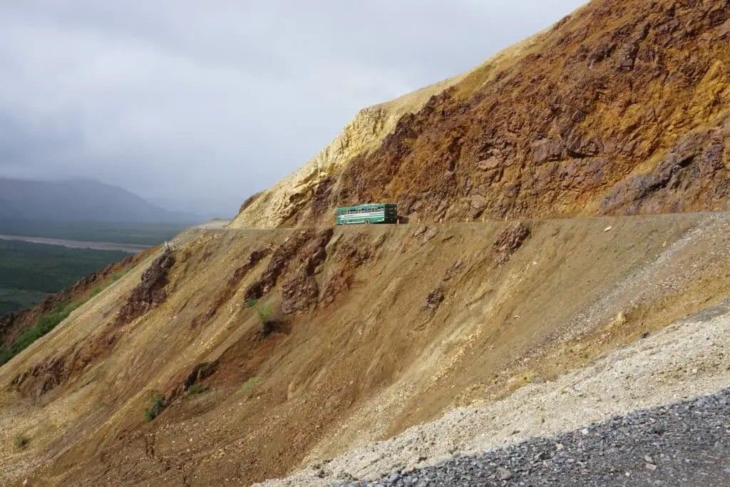 Steep rocky hillside with a small turquoise vehicle on a narrow mountain road under cloudy skies