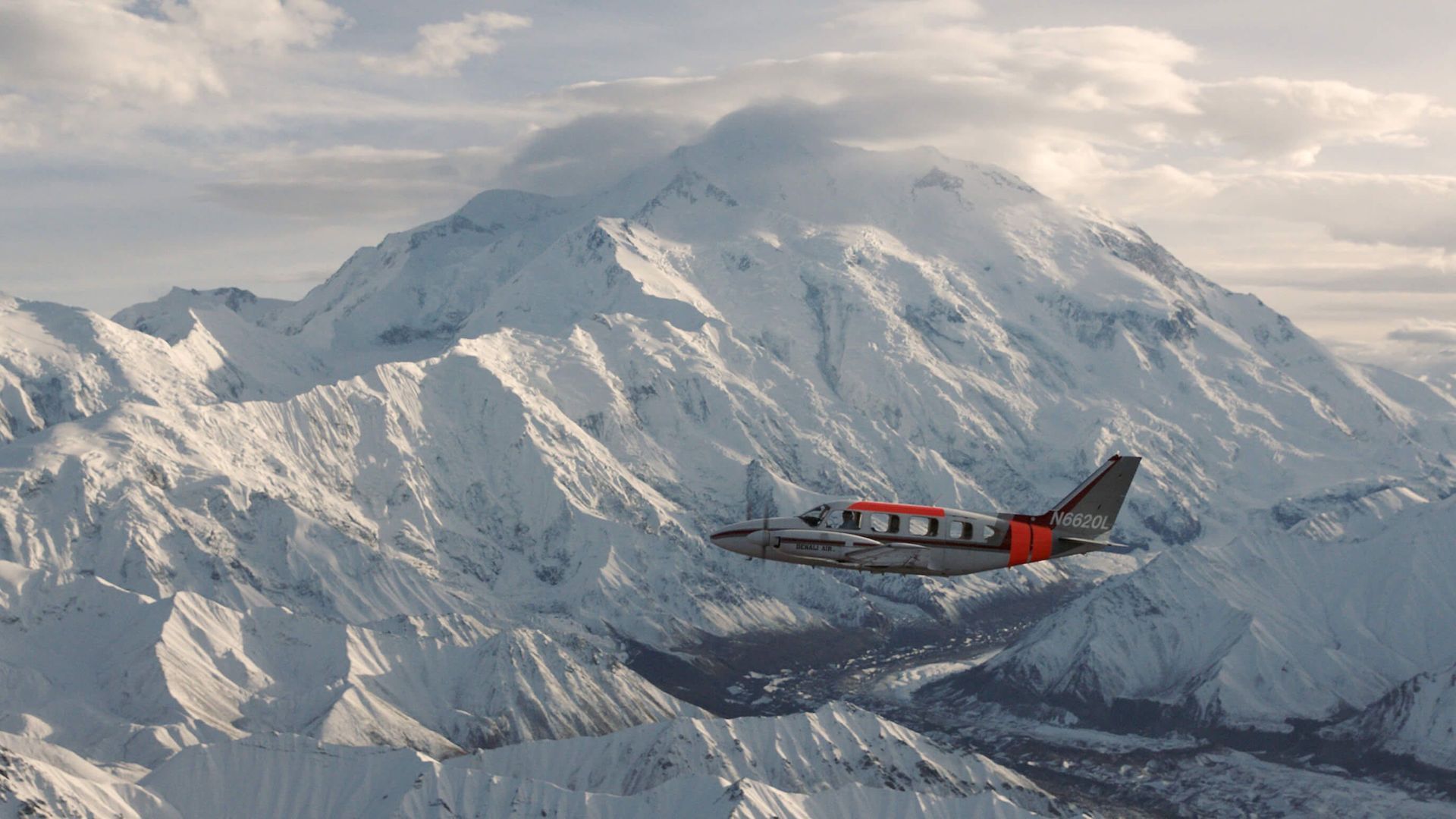 Red-and-black airplane flying beside a snow-covered mountain range under cloudy skies