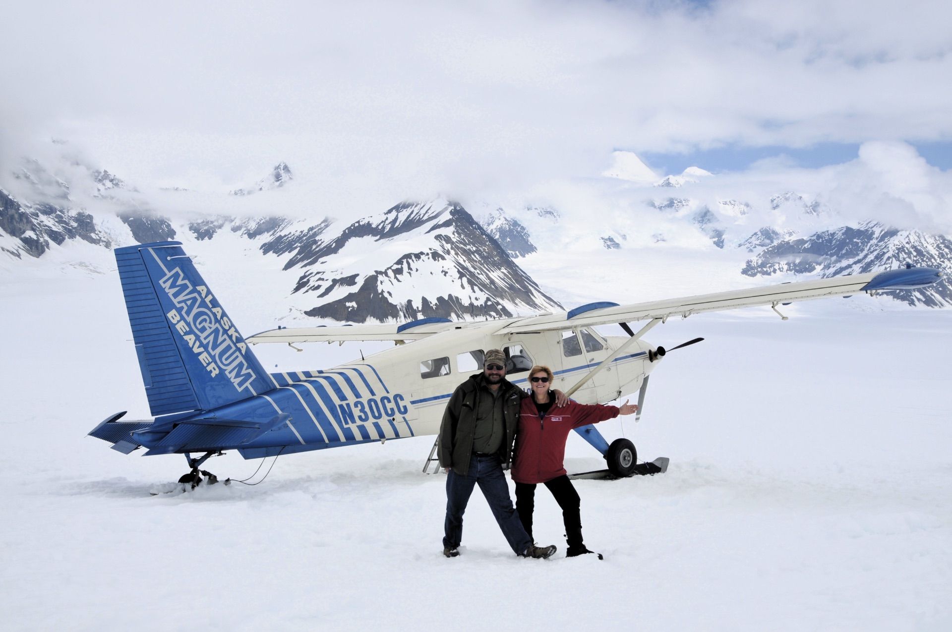 Two people posing beside a blue-and-white ski plane on a snowy mountain airstrip