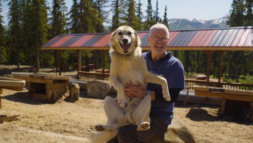 Person holding a smiling dog outdoors in a rustic picnic area with mountains in the background