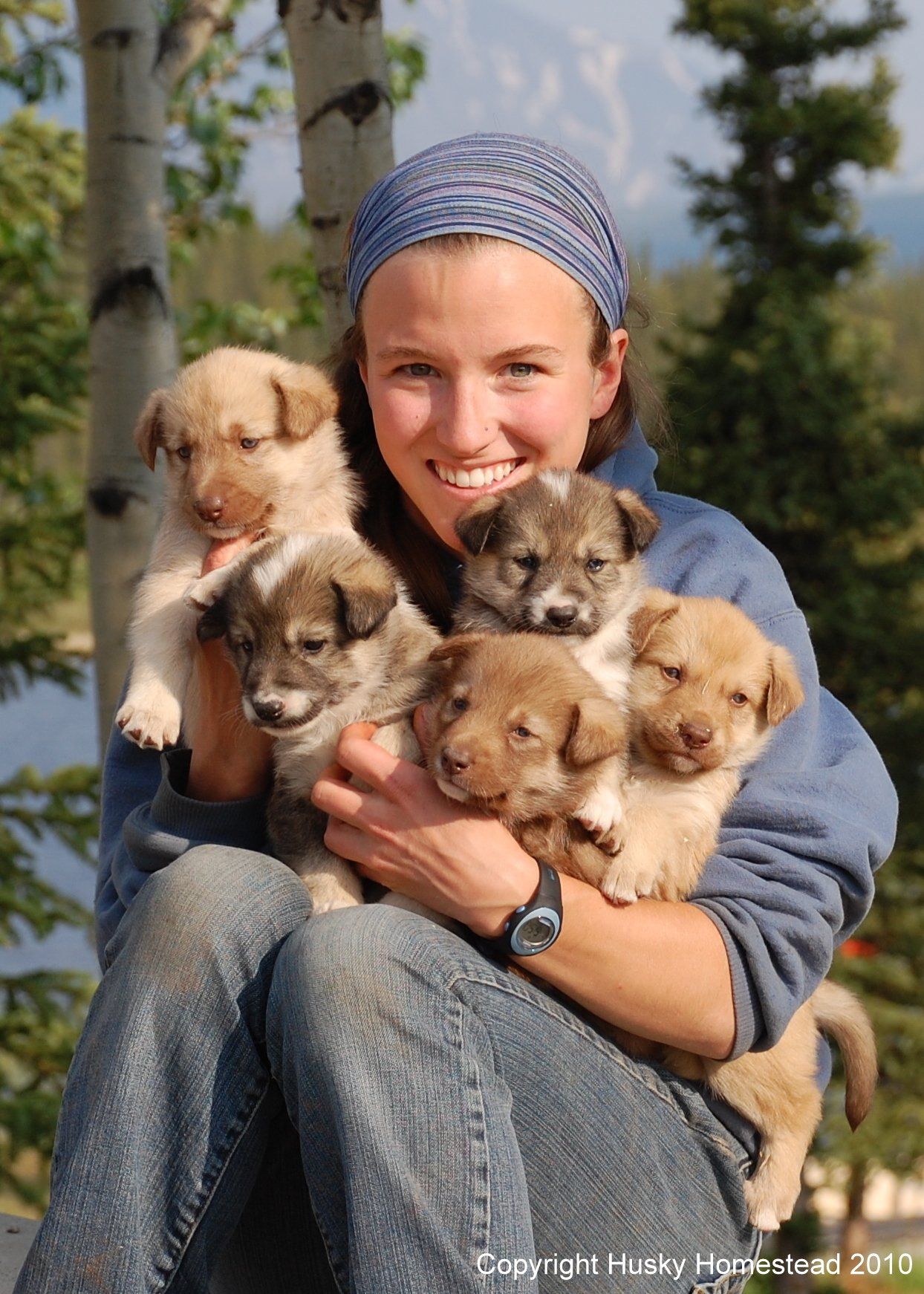 Woman holding five puppies outdoors, smiling in front of trees.