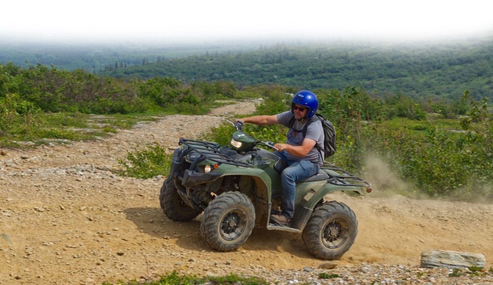 Person riding a black ATV on a rocky dirt trail with forested hills in the background