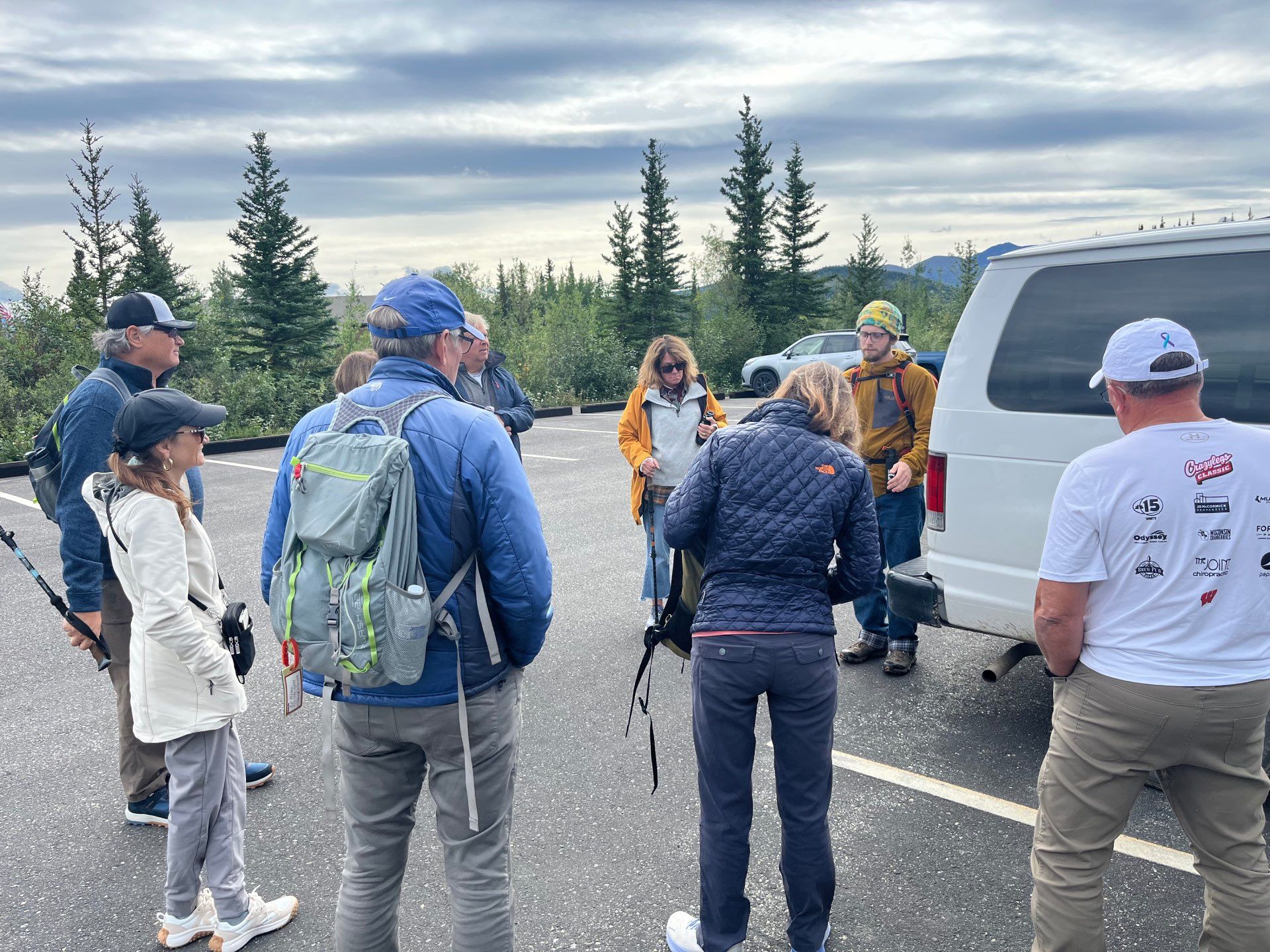 People with backpacks gathered beside a white van in a parking lot under a cloudy sky.