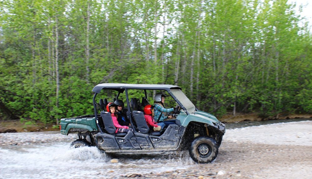 Two people riding in a green off-road buggy through shallow river water in a forest
