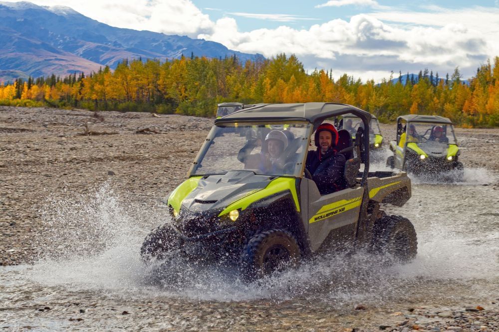 Two off-road vehicles splashing through a rocky river with mountains and autumn trees in the background