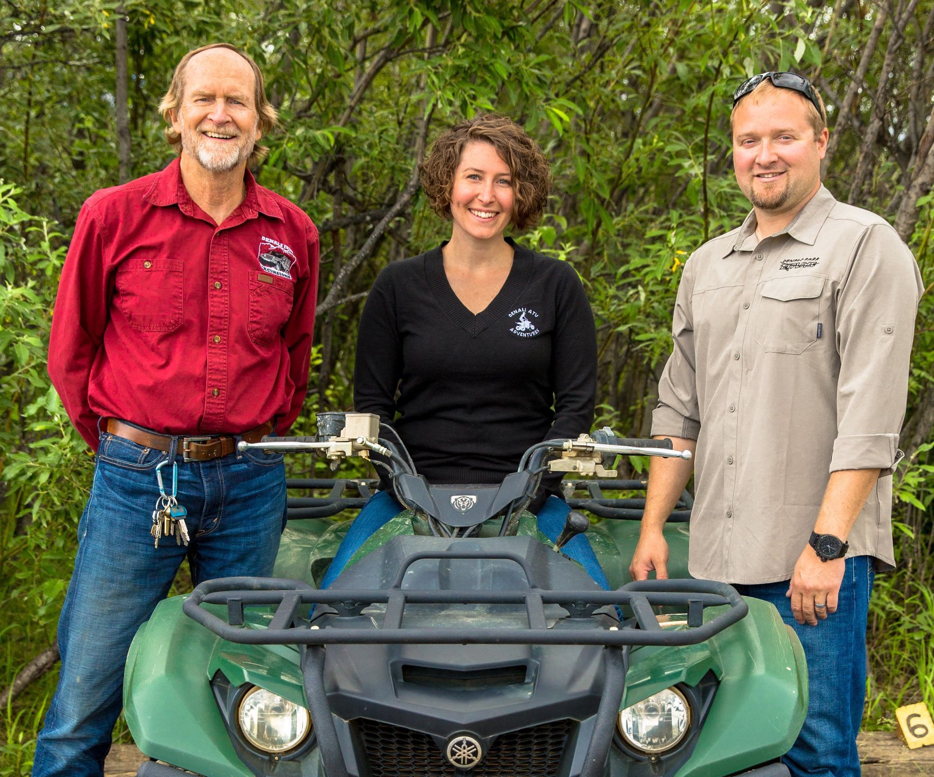 Three people standing behind a green ATV in a wooded outdoor setting, smiling at the camera.