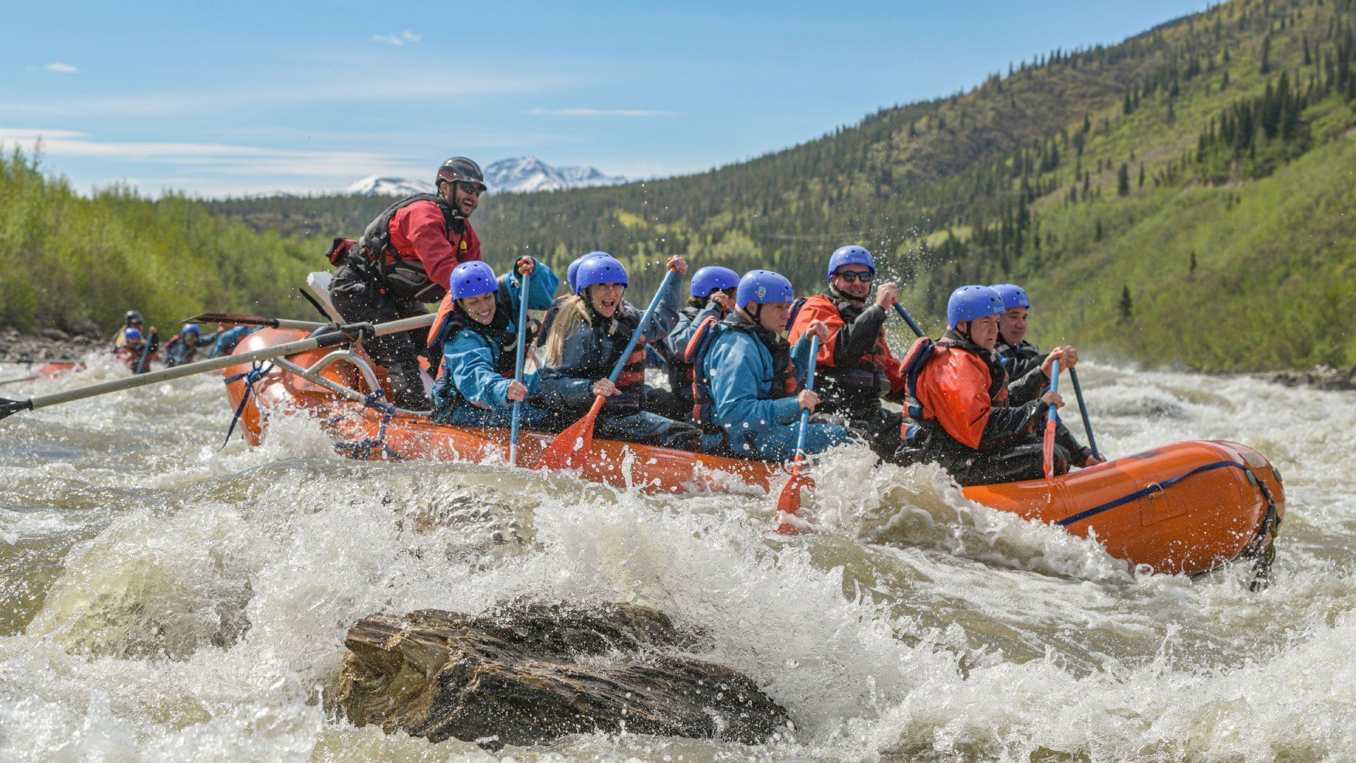 Group whitewater rafting through rapids in an orange raft, wearing helmets and life jackets, with mountains behind