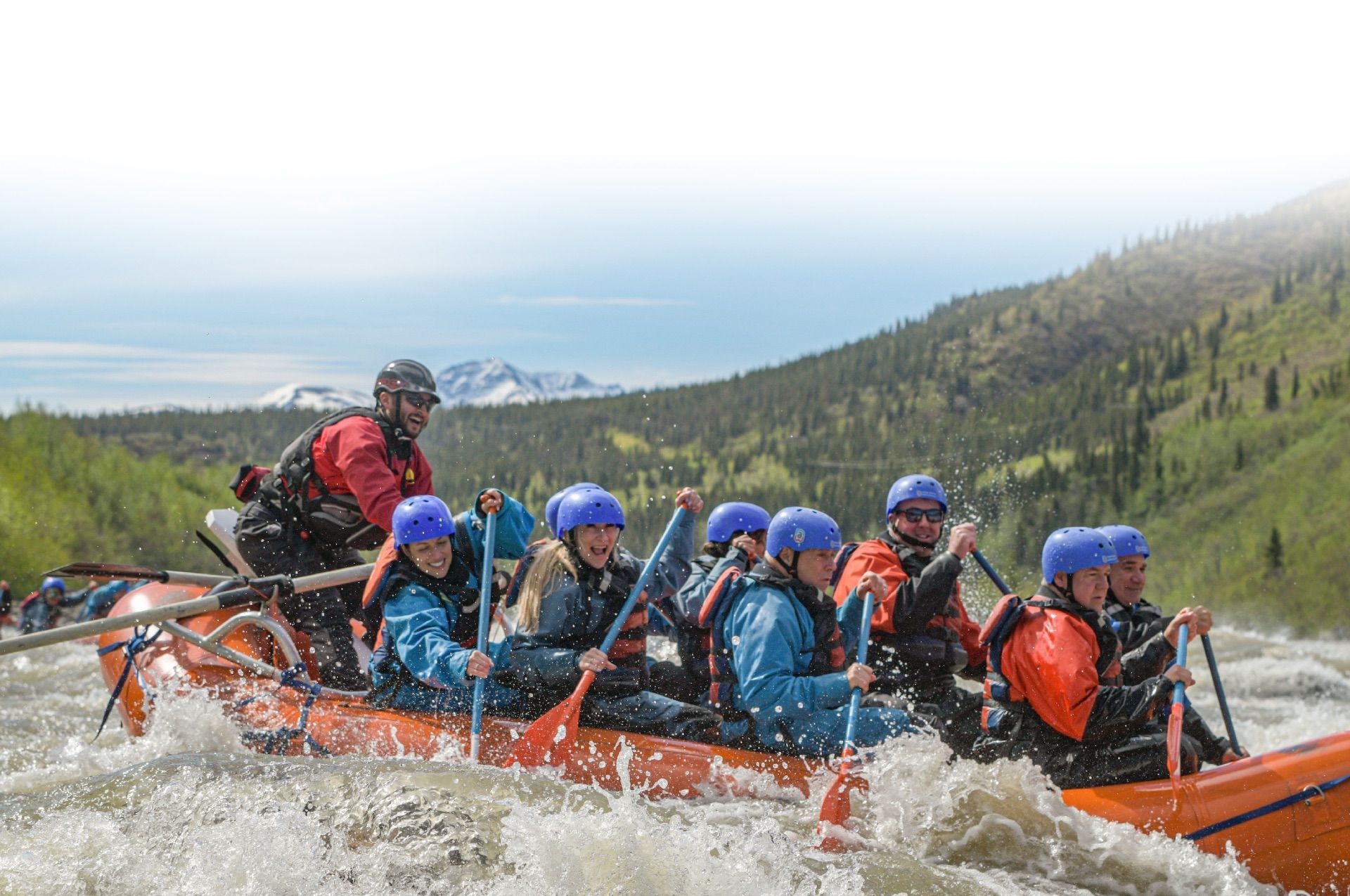 Group whitewater rafting in orange raft with blue helmets on a river, splashing through rapids