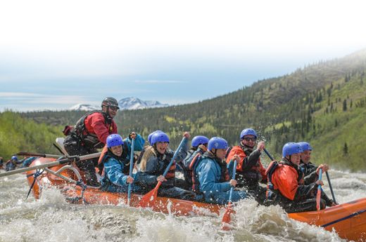 Group rafting in orange boat on choppy river, wearing blue helmets, with forested hills in the background